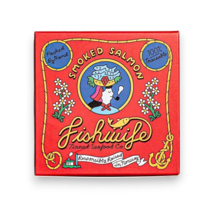 A red square box of FISHWIFE - SMOKED ATLANTIC SALMON is shown. The front showcases vibrant illustrations, including a woman holding a fish on a plate, decorative text, and labels such as "100% Traceable," "Responsibly Raised in America," emphasizing its origins from a family-run cannery that uses sustainable seafood.