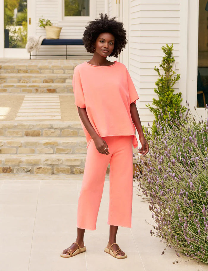 A woman stands outdoors on a stone patio, wearing FRANK & EILEEN'S Catherine Favorite Sweatpant in Wild Strawberry with a pink top and sandals. Steps, greenery, and a light-colored house are visible behind her.