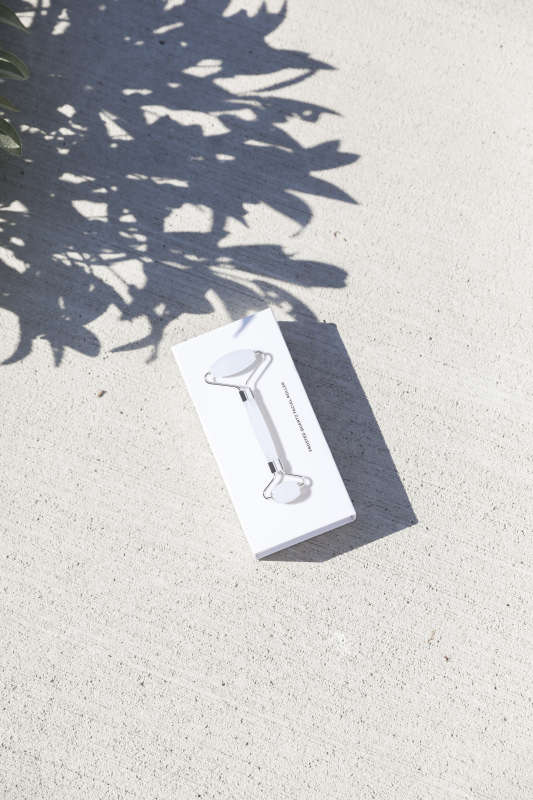A Terra Beauty Bars Frosted Quartz Facial Roller rests on a textured white surface, dappled with leaf shadows. Visible through the boxs transparent section, its designed for facial massage and skin depuffing.