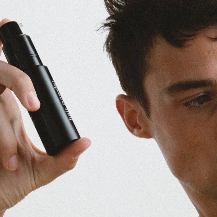 A shirtless person holds a black JACK HENRY – HAIR POWDER bottle, a dry shampoo with smectite, micro algae for growth and shine, and arrowroot to absorb oil—leaving hair clean and matte.