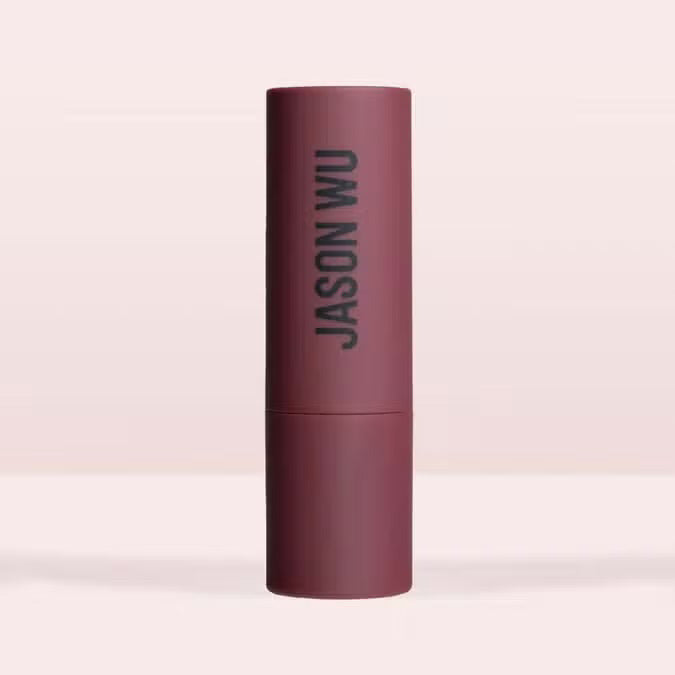 A burgundy JASON WU BEAUTY HOT FLUFF LIP/EYE/CHEEK matte lipstick stands upright with the brand name printed vertically, set against a pale pink background.
