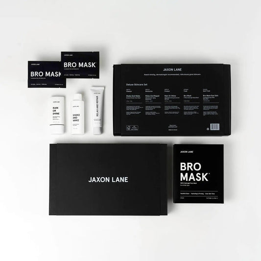 The JAXON LANE - DELUXE SKINCARE SET FOR MEN by JAXON LANE includes the BRO MASK, Rain or Shine sunscreen, Shake and Wake, and two large black boxes, all displayed neatly on a white surface.