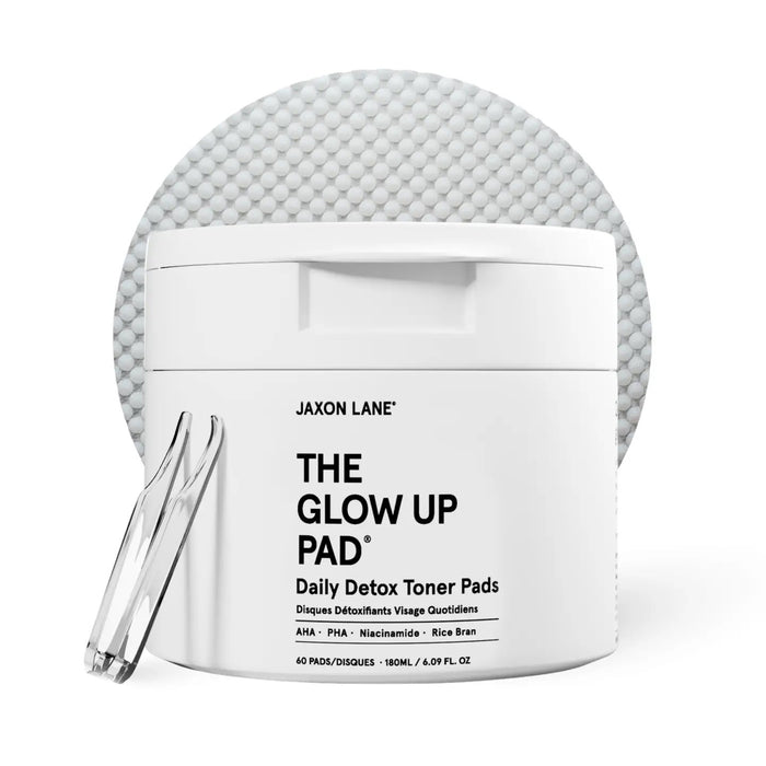 A white jar labeled JAXON LANE - GLOW UP PAD DAILY DETOX TONER PADS sits with a clear tweezer in front of a textured pad. These hydrating, biodegradable pads contain AHA, PHA, niacinamide, and rice bran.