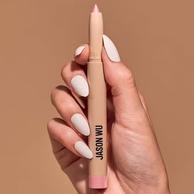 A hand with manicured nails holds a JEWEL STICK EYESHADOW - SPARKLE PEARL by JASON WU BEAUTY, against a light brown background.