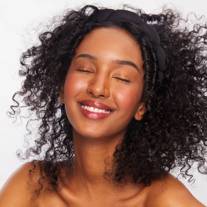 The KITSCH PUFFY HEADBAND, a black and eco-friendly accessory with a padded, segmented design, is artfully displayed against a plain white backdrop. A small tag emphasizing its sustainable nature is attached to one side.