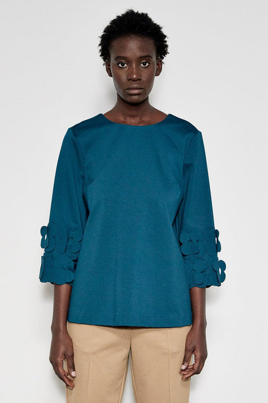 A person wears the MEIMEIJ 3/4 Sleeve Back To Front Top with Lasercut Flower Detail in teal and beige pants, standing against a plain white background.
