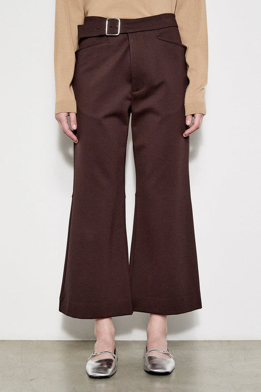 A person models the MEIMEIJ Crop Ponte Pant with Belt, shown in brown with pockets, paired with a beige top and silver flats. Only the lower half is visible against a plain white wall and concrete floor.