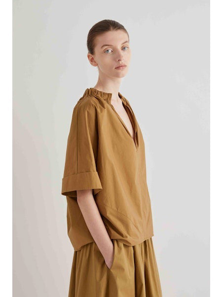 A person wearing the MEIMEIJ Gathered Neck Blouse in Off White by MEIMEIJ stands against a plain light background, looking calmly at the camera with hands in pockets.