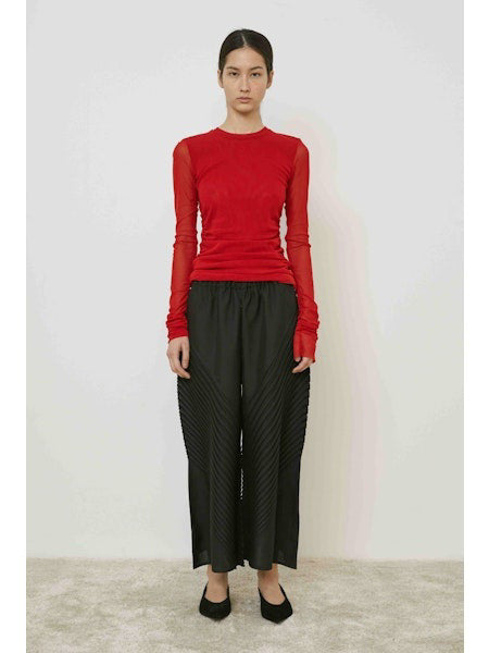 A woman with long dark hair in a low ponytail stands in profile against a plain light background, wearing the MEIMEIJ MESH LAYERING TOP by MEIMEIJ in red and a black skirt for a minimalist Italian fashion look.