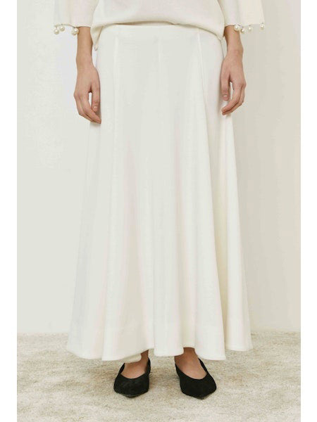 A person wears the MEIMEIJ - SKIRT, a long, flowing minimalist piece by MEIMEIJ, styled with a white top with pearl-detailed sleeves and black flats, standing on a light carpet.