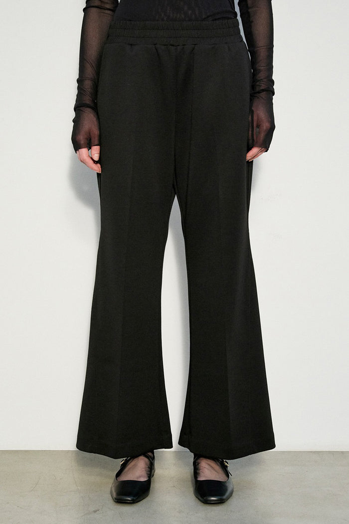 A person stands against a white wall wearing MEIMEIJ - STRIGHT LEG PULL ON PONTE PANT by MEIMEIJ with a sheer black long-sleeve top and black shoes. Only the lower body is visible; the upper body and head are not shown.