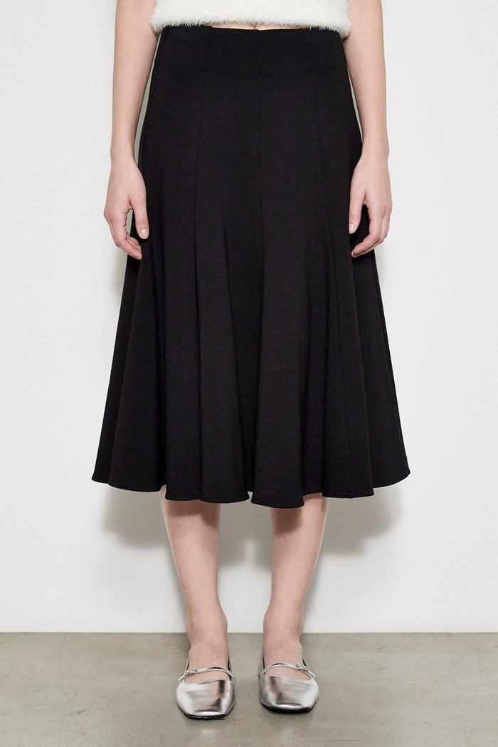 A person models the MEIMEIJ SWING SKIRT WITH LACE BACK in black, paired with silver flats, standing against a white wall. The photo is cropped at the waist and does not reveal the face.