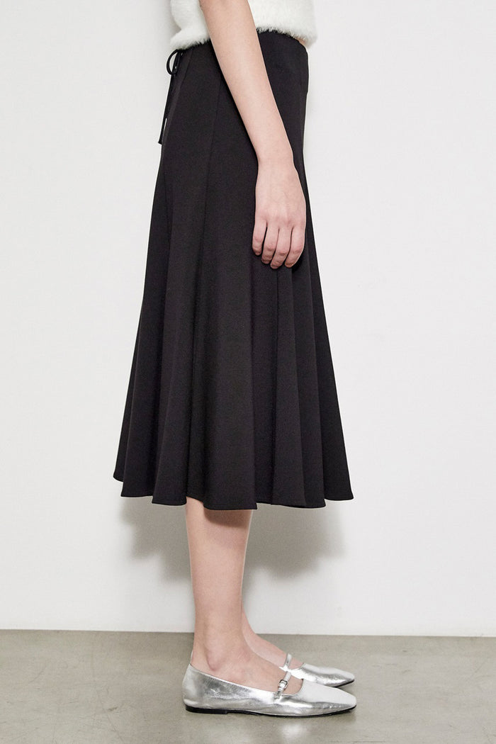 A person models the MEIMEIJ SWING SKIRT WITH LACE BACK in black, paired with silver flats, standing against a white wall. The photo is cropped at the waist and does not reveal the face.