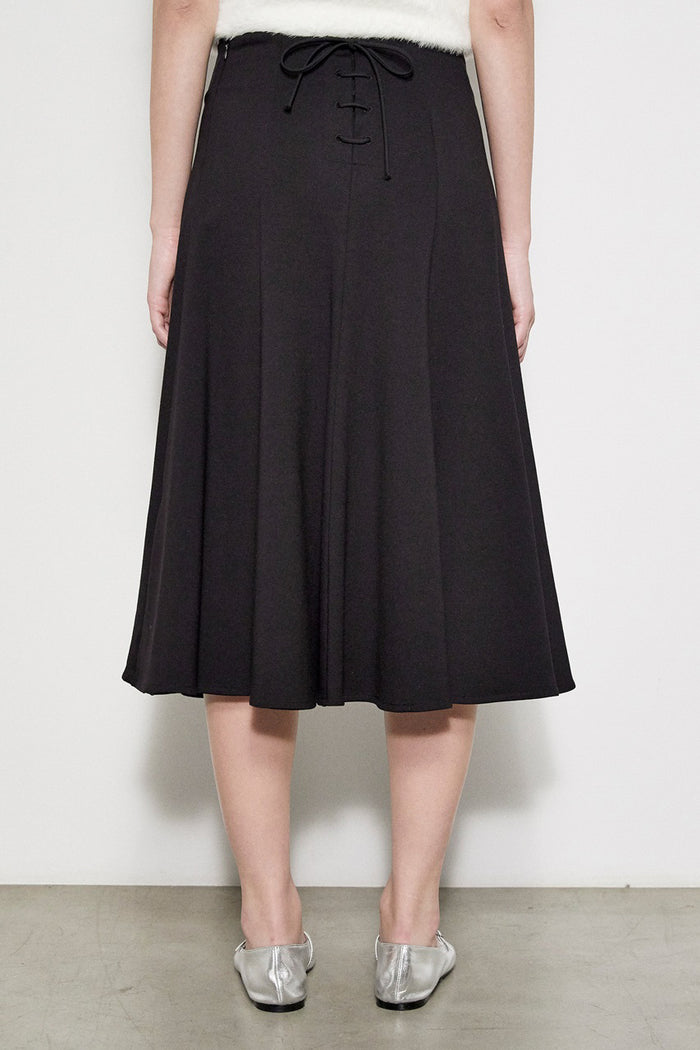 A person models the MEIMEIJ SWING SKIRT WITH LACE BACK in black, paired with silver flats, standing against a white wall. The photo is cropped at the waist and does not reveal the face.