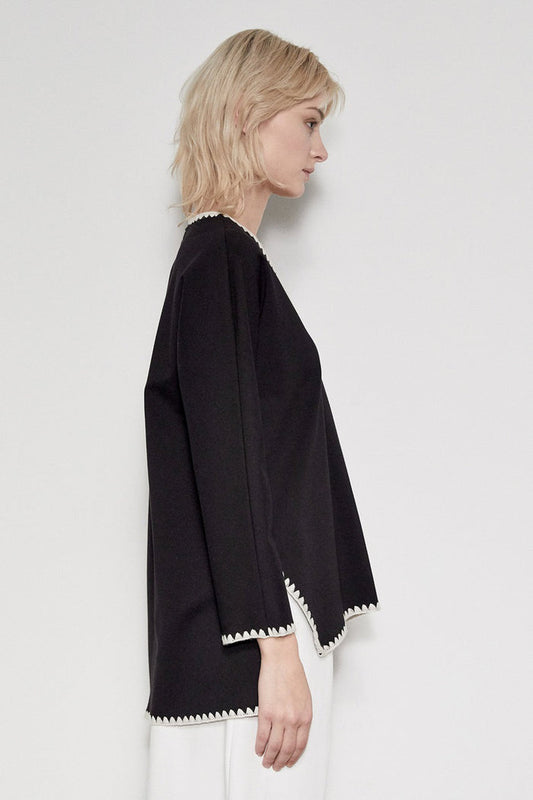 A person with light blonde hair stands sideways against a white background, wearing the MEIMEIJ V Neck Hi Low Top with embroidered contrast stitch detail by MEIMEIJ, paired with white pants.
