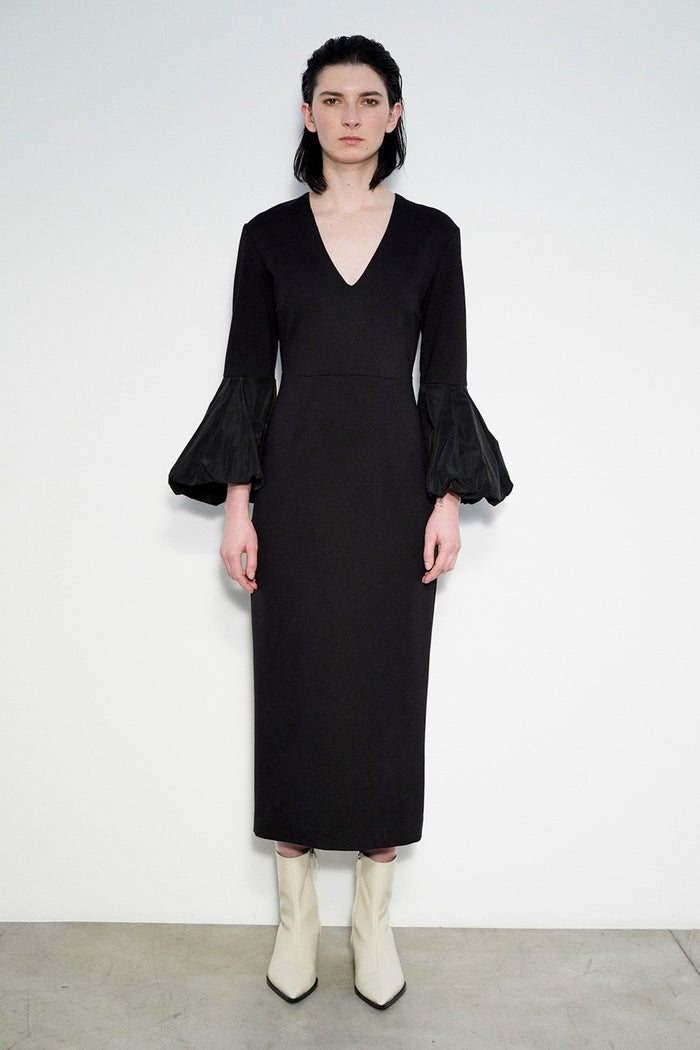 A person stands against a white wall, wearing the MEIMEIJ V Neck Midi Dress with puff sleeves and off-white pointed-toe ankle boots. Their straight dark hair and Italian craftsmanship complete the elegant look.