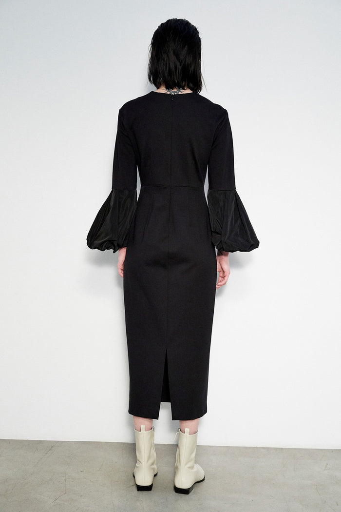 A person stands against a white wall, wearing the MEIMEIJ V Neck Midi Dress with puff sleeves and off-white pointed-toe ankle boots. Their straight dark hair and Italian craftsmanship complete the elegant look.