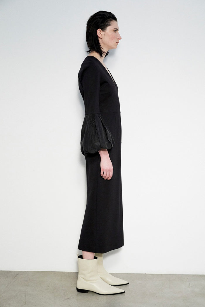 A person stands against a white wall, wearing the MEIMEIJ V Neck Midi Dress with puff sleeves and off-white pointed-toe ankle boots. Their straight dark hair and Italian craftsmanship complete the elegant look.