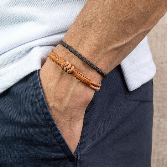 A person with a hand in their navy blue pants pocket wears two bracelets—one thin and dark, the other a thicker MAKARLA Men's Knot Leather Bracelet Adjustable in Caramel—set against a light background.