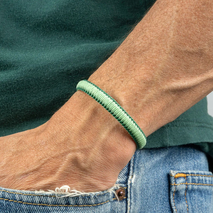 A close-up of a person's wrist wearing the MAKARLA MEN'S REVERSIBLE SURFER BRACELET in mint/hunter green. The person is dressed in a green shirt and blue jeans with a small tear near the pocket.