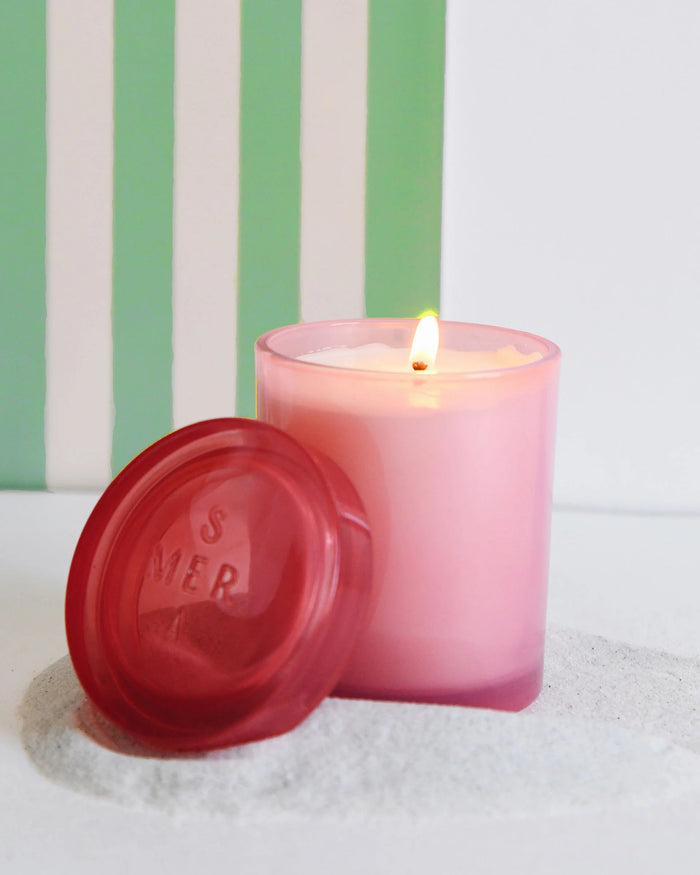 The lit MERSEA - CABANA FLOWER JOLIE JAR CANDLE, infused with ylang ylang, graces a translucent pink jar with its matching lid on a white surface. Green and white stripes in the backdrop enhance its serene charm, while the embossed lettering on the lid adds sophistication.