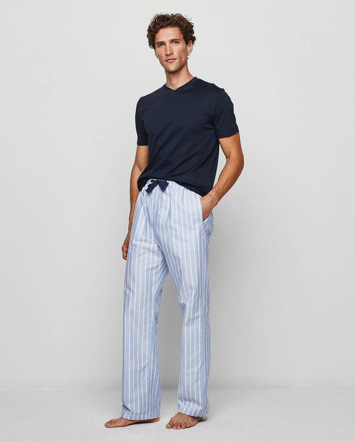 A man stands barefoot against a plain background, wearing the MIRTO 1956 - OXFORD PAJAMA SET by MIRTO 1956, featuring a dark blue top and light blue and white striped cotton pants with a comfortable drawstring waist.