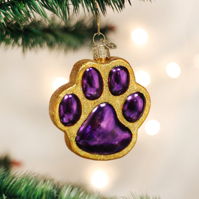 A shiny Christmas tree ornament shaped like a paw print, with gold glitter edges and glossy purple pads, hangs from a green tree branch. Blurred white holiday lights glow in the background.