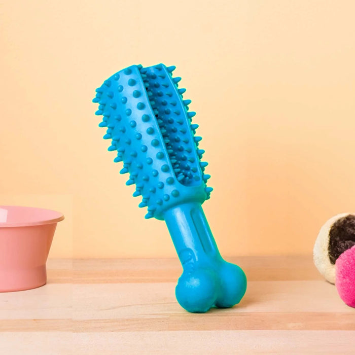 A blue Pet Dental Chew Toy, bone-shaped with textured spikes by Oxyfresh, stands upright on a wooden surface. Its flanked by a pink bowl on the left and a plush toy on the right, all set against a light beige background.