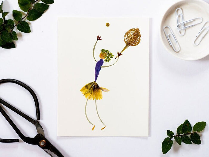 The PETAL PEOPLE PRESS - GAME, SET, MATCH CARD by PETAL PEOPLE PRESS features a whimsical tennis figure crafted from pressed flowers and leaves. It sports a purple upper body, yellow skirt, green leafy head, and holds a plant material racket reaching toward a Trout Lily-like ball.