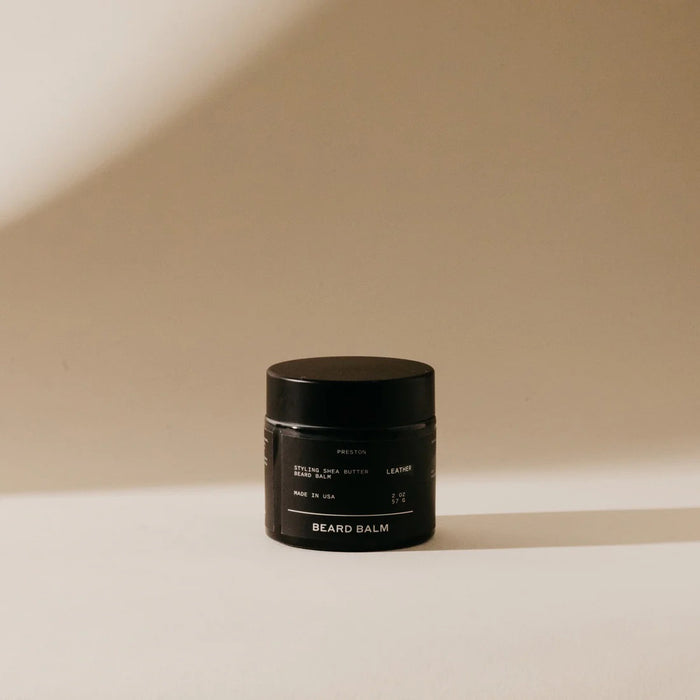 A small black jar labeled "PRESTON - BEARD BALM" by PRESTON sits on a light tan surface, its shea butter-infused formula ideal for beard shaping and daily grooming, highlighted by the minimalist background and soft shadow.