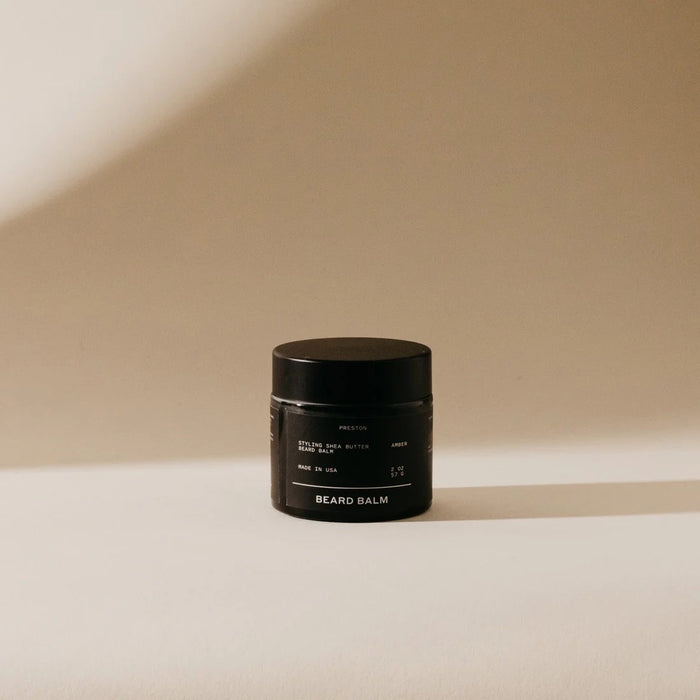 A small black jar labeled "PRESTON - BEARD BALM" by PRESTON sits on a light tan surface, its shea butter-infused formula ideal for beard shaping and daily grooming, highlighted by the minimalist background and soft shadow.