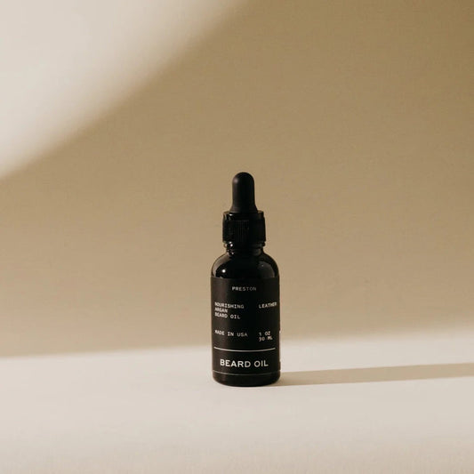 A small dark glass bottle of PRESTON - BEARD OIL by PRESTON, with a dropper cap, sits on a light surface casting a soft shadow. The label displays "PRESTON - BEARD OIL" along with product details in white text.