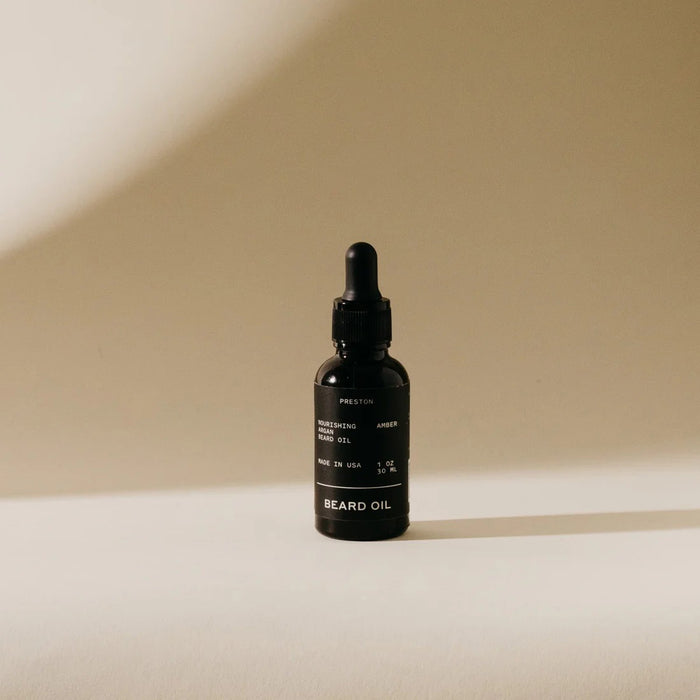 A small dark glass bottle of PRESTON - BEARD OIL by PRESTON, with a dropper cap, sits on a light surface casting a soft shadow. The label displays "PRESTON - BEARD OIL" along with product details in white text.