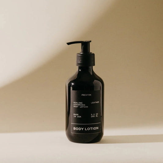 A dark bottle of PRESTON - BODY LOTION 9.5OZ with a pump dispenser rests on a light surface in soft lighting. The label features the PRESTON brand and details about plant-based moisturizers in white text.