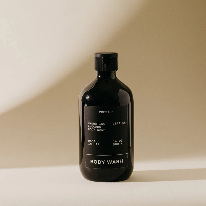 A 16 oz PRESTON - BODY WASH by PRESTON with a black label, white text, and black cap sits on a light surface against a beige background. This hydrating, sulfate-free body wash comes in a dark bottle.