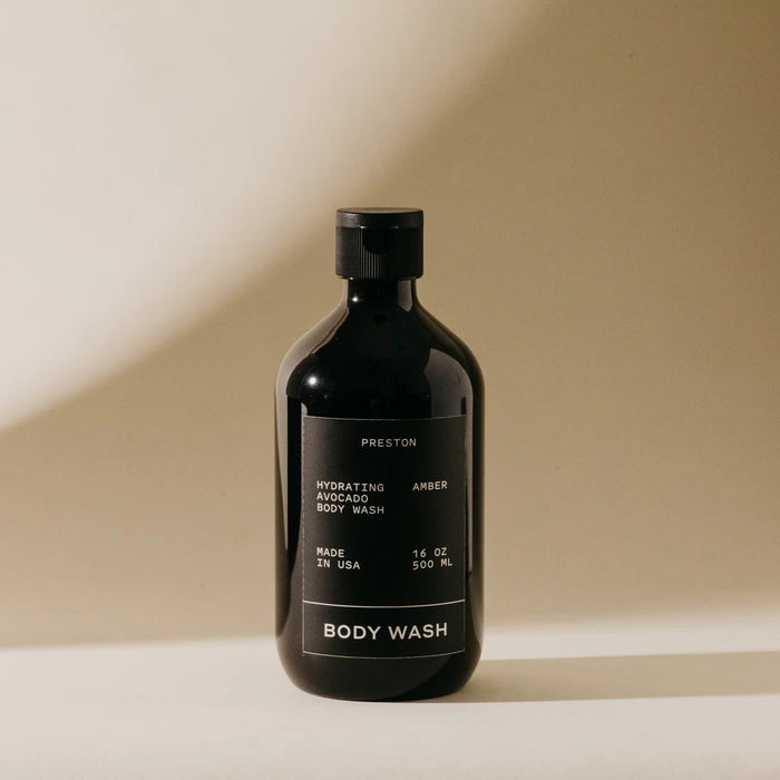 A 16 oz PRESTON - BODY WASH by PRESTON with a black label, white text, and black cap sits on a light surface against a beige background. This hydrating, sulfate-free body wash comes in a dark bottle.