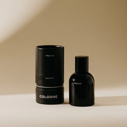 A black cylindrical PRESTON - COLOGNE 1.7OZ bottle stands beside its matching packaging, both labeled "PRESTON," on a beige background, evoking the subtle scent inspired by essential oil cologne blends.