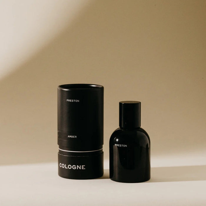 A black cylindrical PRESTON - COLOGNE 1.7OZ bottle stands beside its matching packaging, both labeled "PRESTON," on a beige background, evoking the subtle scent inspired by essential oil cologne blends.