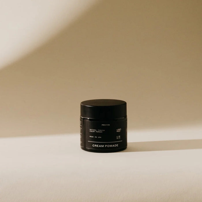 A black jar labeled "PRESTON - CREAM POMADE UNSCENTED" rests on a beige surface with a soft shadow, highlighting its matte finish and light hold formula by PRESTON, set against a neutral background.
