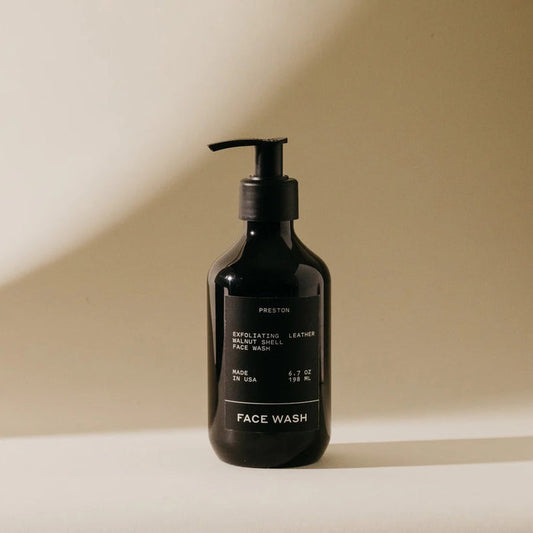 A black pump bottle labeled "PRESTON - FACE WASH" by PRESTON sits on a beige surface in soft, natural light. The minimalist label notes it’s a resurfacing exfoliating cleanser with walnut shells to unclog pores, made in the USA.