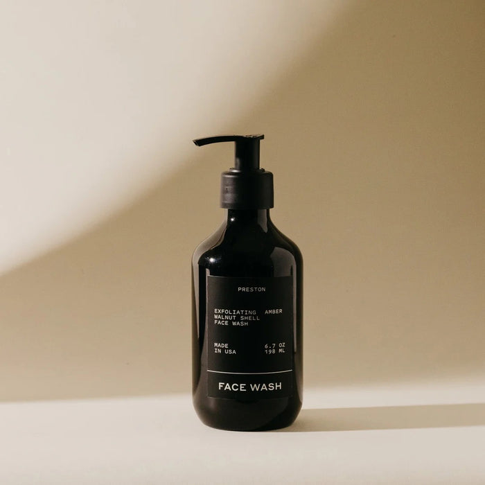 A black pump bottle labeled "PRESTON - FACE WASH" by PRESTON sits on a beige surface in soft, natural light. The minimalist label notes it’s a resurfacing exfoliating cleanser with walnut shells to unclog pores, made in the USA.