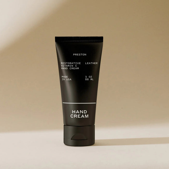 A black PRESTON HAND CREAM 3OZ tube stands upright on a neutral surface with a beige background. Made in the USA, this cream deeply hydrates and nourishes hands with vitamin C benefits.