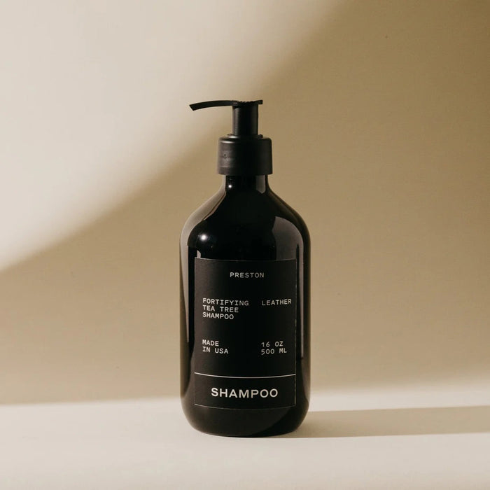 A black pump bottle labeled "PRESTON - SHAMPOO" sits on a beige surface with soft lighting. This sulfate-free shampoo, made in the USA by PRESTON, contains tea tree oil and leather scent and comes in a 16 oz (500 ml) size.