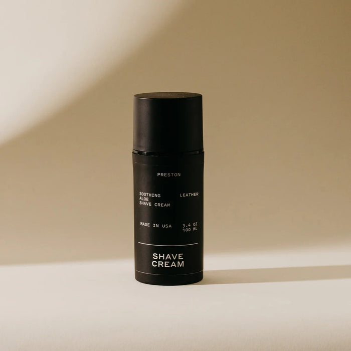 A black cylindrical bottle of PRESTON - SHAVE CREAM stands on a light beige surface with a soft shadow, featuring minimalist packaging with white text that highlights its cooling formula infused with soothing aloe vera.