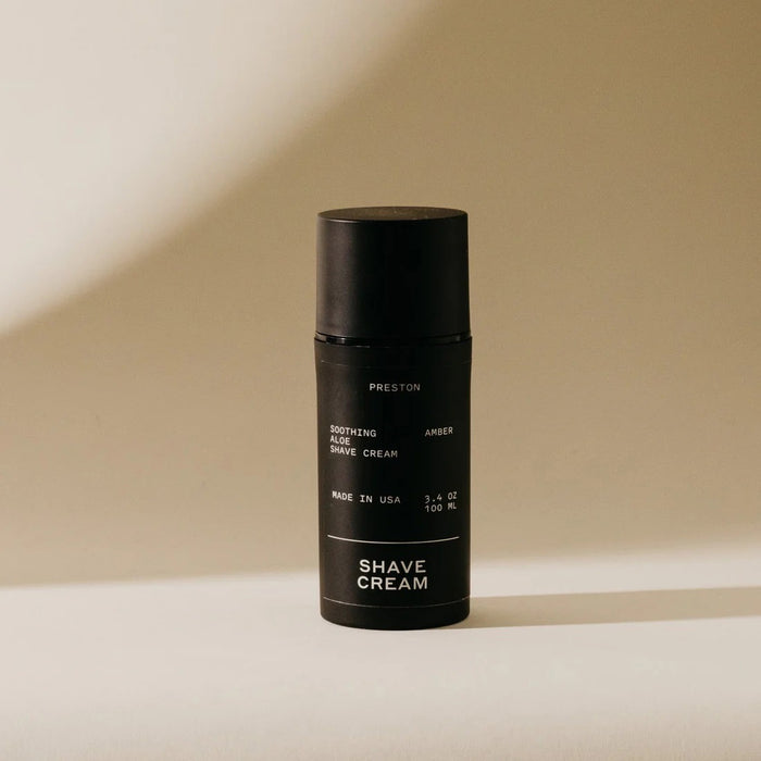 A black cylindrical bottle of PRESTON - SHAVE CREAM stands on a light beige surface with a soft shadow, featuring minimalist packaging with white text that highlights its cooling formula infused with soothing aloe vera.