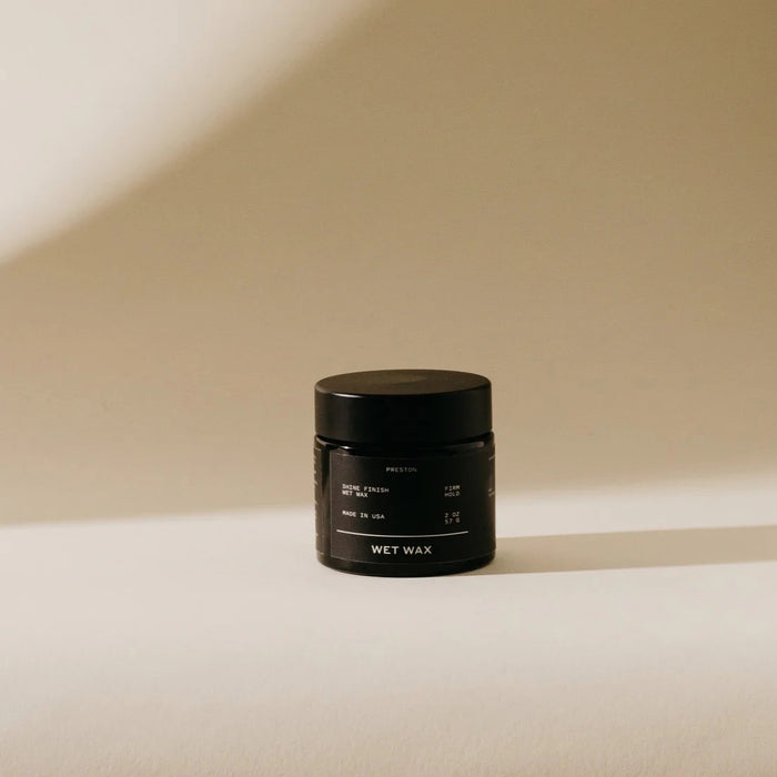 A small black jar labeled "PRESTON - WET WAX," a vegan hair product by PRESTON, sits on a light surface with a soft beige background and its shadow cast to the right.