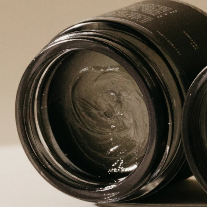 A small black jar labeled "PRESTON - WET WAX," a vegan hair product by PRESTON, sits on a light surface with a soft beige background and its shadow cast to the right.