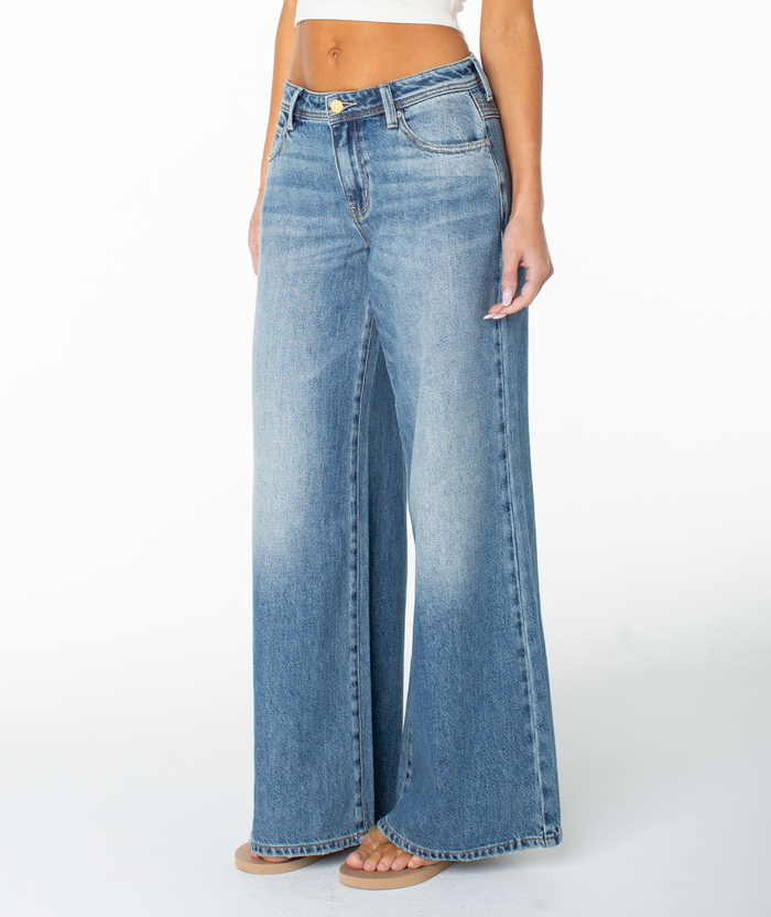 Someone models the Roxy Refuge Retro Pant, featuring a high waist, wide leg, slight faded wash, and front pockets. Styled with a white crop top and barefoot, they stand against a plain white background.