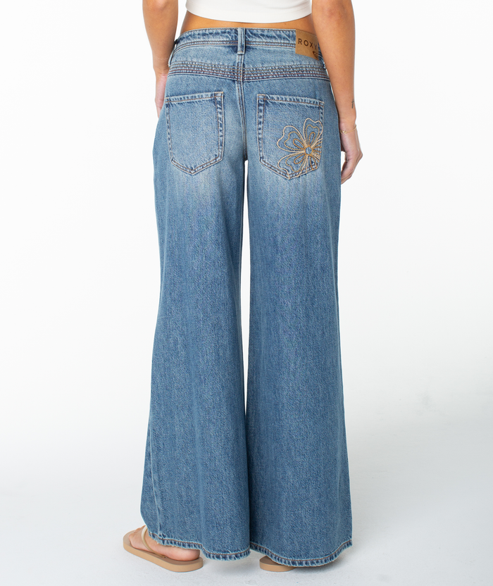 A person wearing Roxy Refuge Retro Pant jeans with decorative stitching on one back pocket, paired with a white crop top and sandals, is shown from the back against a plain white background.
