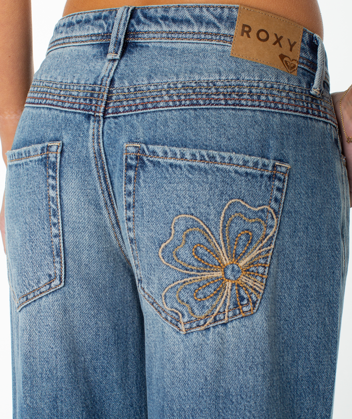 Close-up of the back of a person wearing blue low waisted Refuge Retro Pant jeans by Roxy with a flower embroidered on one pocket and a brown Roxy label on the waistband.